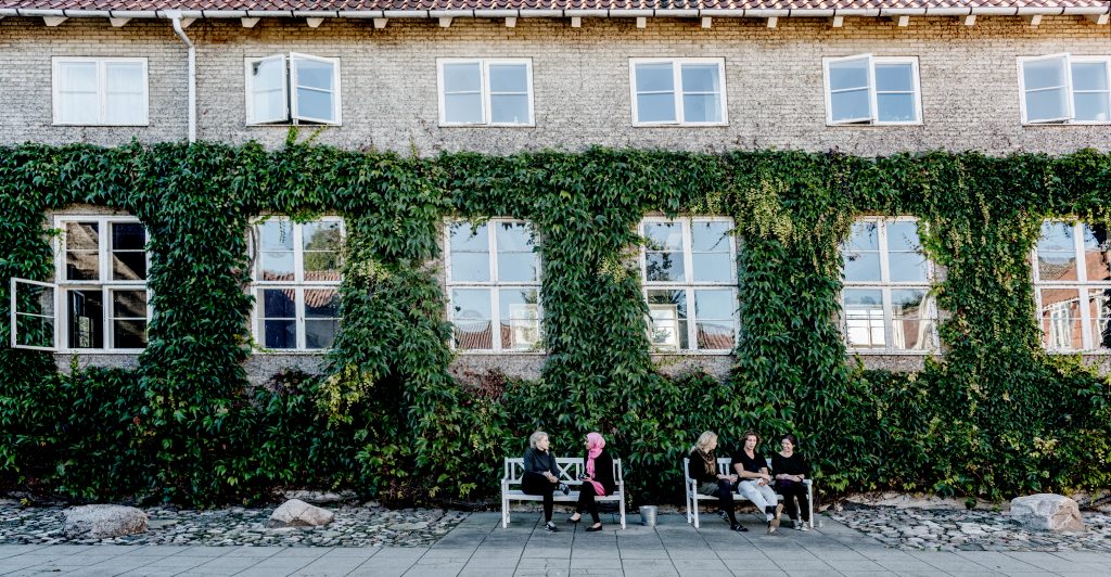 Fem mennesker sidder på to bænke og snakker foran en stor bygning med grøn plante, der vokser op ad bygningen.